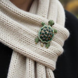 May include: A gold-toned turtle brooch with green rhinestones is pinned to a cream-colored knit scarf. The turtle's shell is detailed with dark green stones, and its legs and head are adorned with smaller green rhinestones.