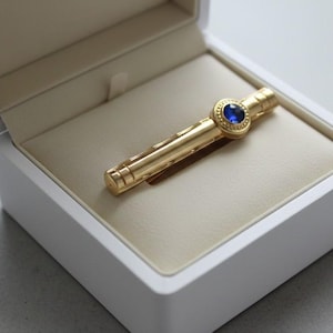 May include: A gold-colored tie clip with a round, blue gemstone set in the center. The tie clip is in a white box with a beige interior. The box is open, displaying the tie clip.