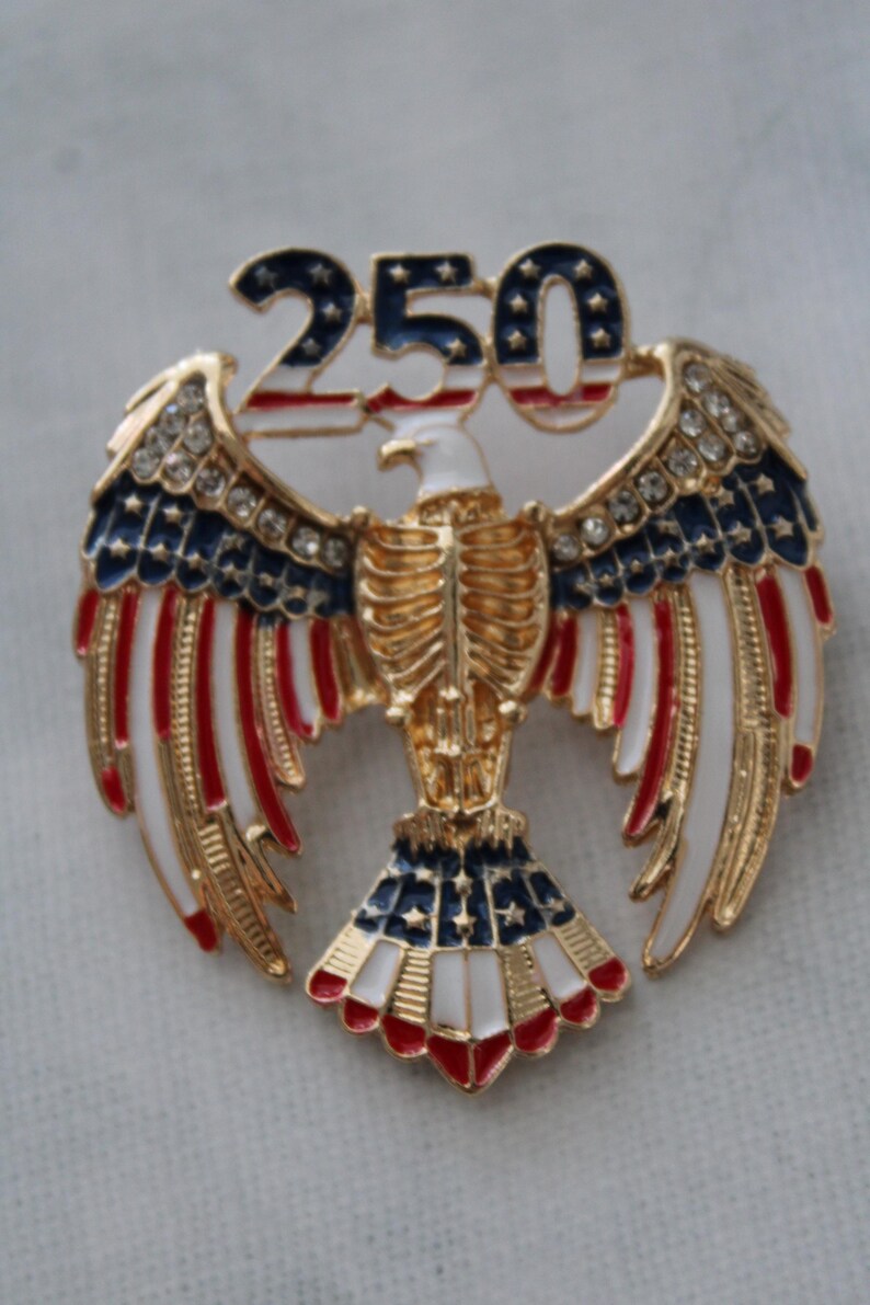 May include: A gold-toned eagle pin with outstretched wings, featuring red, white, and blue enamel accents. The number "250" is displayed above the eagle, also in red, white, and blue. The pin is embellished with small, clear rhinestones.