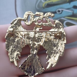 May include: Gold-toned metal brooch in the shape of a stylized bird with outstretched wings. The brooch has a textured surface and a pin closure on the back. The word "OFF" is at the top of the brooch.