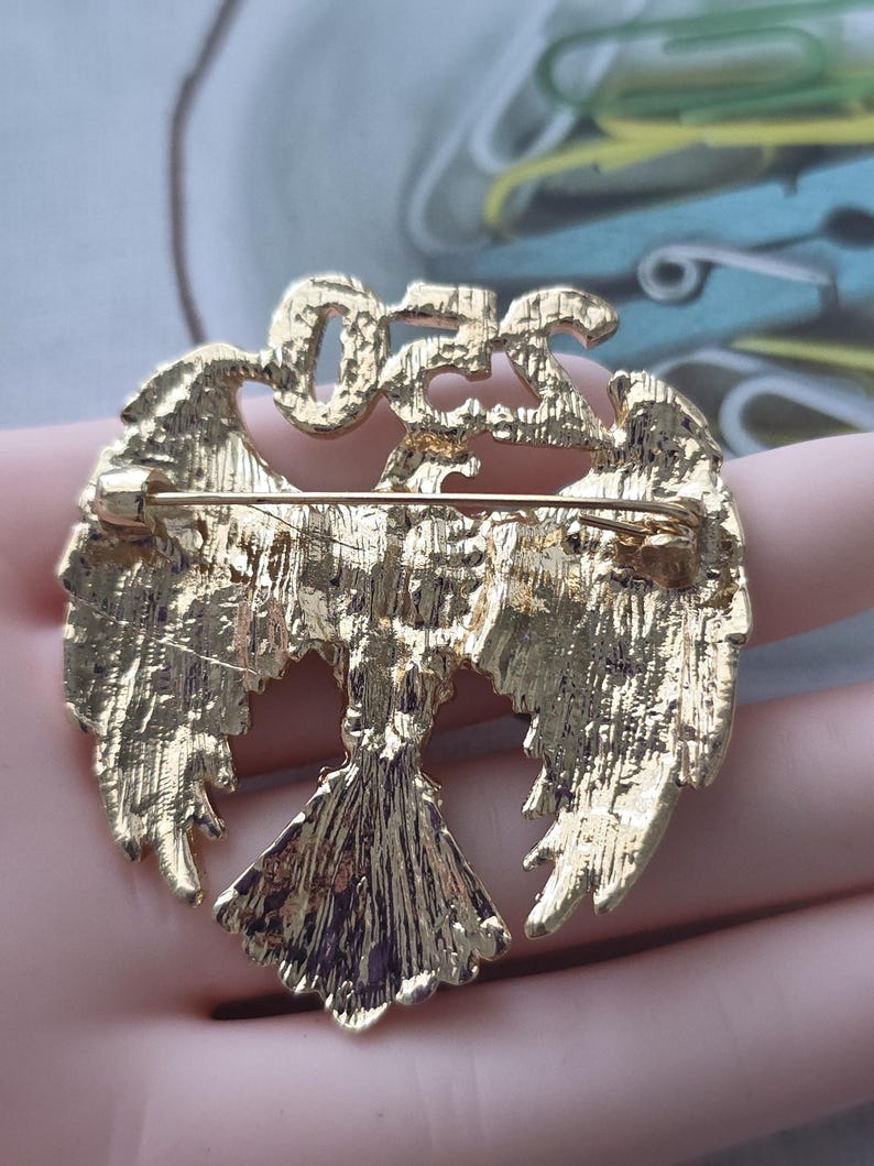 May include: Gold-toned metal brooch in the shape of a stylized bird with outstretched wings. The brooch has a textured surface and a pin closure on the back. The word "OFF" is at the top of the brooch.