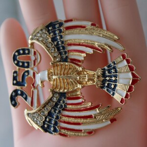 May include: A gold-toned eagle brooch with red, white, and blue enamel detailing. The brooch features the number "250" and is adorned with rhinestones. The eagle's wings and tail display the colors of the American flag.