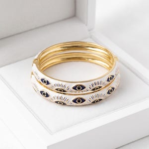 May include: Gold-tone bangles with white enamel and blue eye designs. The bracelets are displayed in a white box. Each bangle features a repeating pattern of blue eyes with gold and black accents.