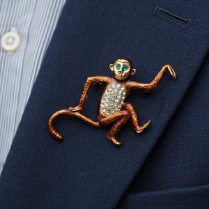 May include: A gold-toned monkey brooch with a red-brown finish, embellished with clear crystals on the body and green gems for the eyes. The brooch is pinned to a navy blue blazer.