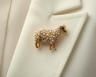 Op de afbeelding: Een goudkleurige schapenbroche, versierd met imitatieparels, is vastgemaakt aan een witte blazer. Het lichaam van het schaap is bedekt met ronde parels, met een gedetailleerde kop en poten. De broche voegt een vleugje elegantie toe aan het kledingstuk.