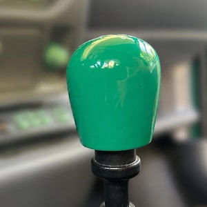Könnte beinhalten: Ein glatter, glänzender, hellgrüner Schaltknauf mit abgerundeter Oberseite, montiert auf einem schwarzen zylindrischen Schaft, der mit einer schwarzen gummierten Basis verbunden ist, im Innenraum eines Fahrzeugs mit unscharfem Armaturenbrett.