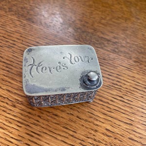 Op de afbeelding: Een kleine, rechthoekige zilveren doos met de inscriptie "Here's Love" op het deksel. De doos heeft een decoratieve rand en een kleine knop aan de zijkant. De doos ligt op een houten oppervlak.