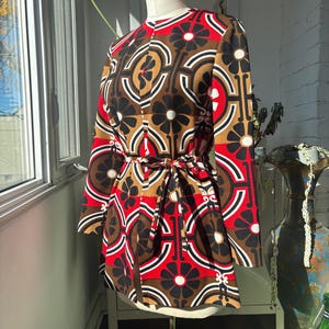 May include: A long-sleeved jacket featuring a bold geometric and floral pattern in red, black, white, and tan. The garment has a round neckline, a front button closure, and a matching fabric tie belt at the waist, displayed on a mannequin.