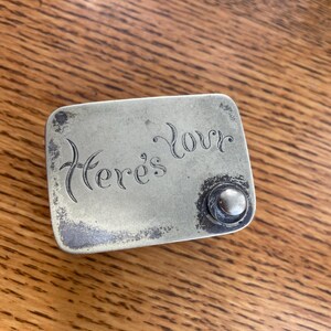 Op de afbeelding: Een rechthoekige, aangetaste metalen doos met de woorden "Here's Love" gegraveerd op de bovenkant. Aan de rechterkant zit een kleine, ronde knop. De doos ligt op een houten oppervlak.