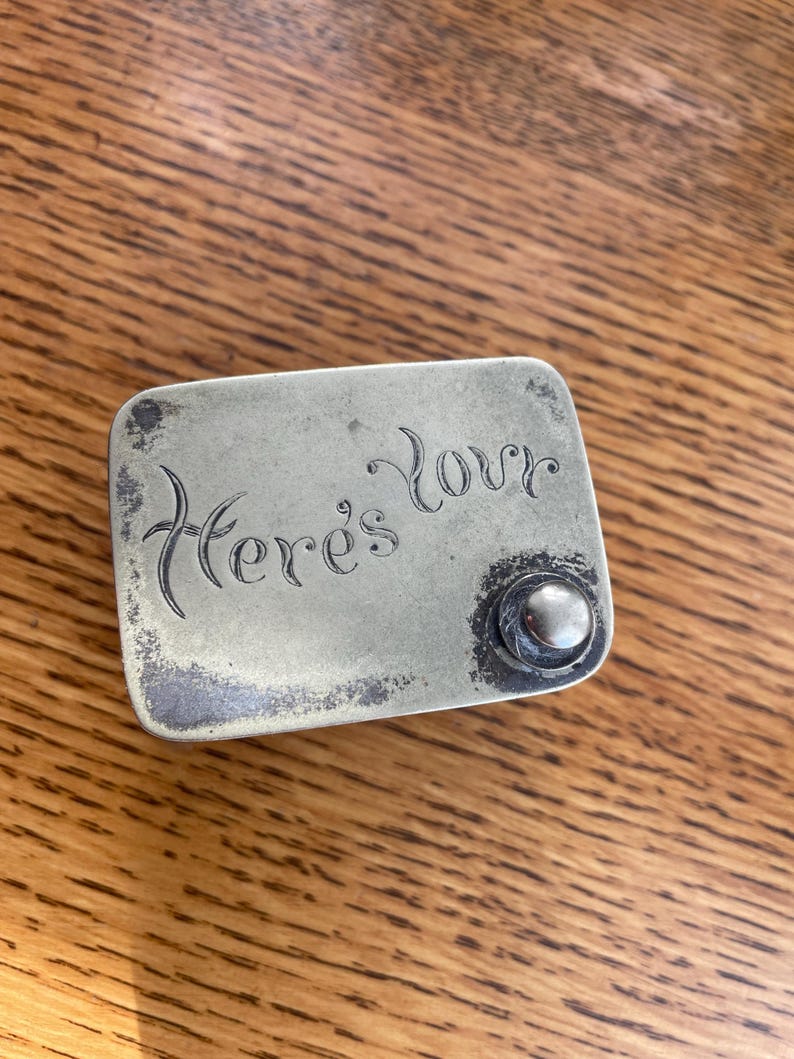 Op de afbeelding: Een rechthoekige, aangetaste metalen doos met de woorden "Here's Love" gegraveerd op de bovenkant. Aan de rechterkant zit een kleine, ronde knop. De doos ligt op een houten oppervlak.