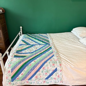 Puede incluir: Una cama blanca con un edredón blanco con estampado floral y rayas verdes, azules y rosas. La cama está contra una pared verde.