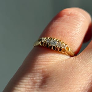 Puede incluir: Primer plano de un anillo de oro con tres piedras preciosas transparentes. El anillo tiene un diseño calado detallado a lo largo de la banda. El anillo está en un dedo, con el fondo borroso.