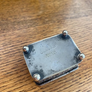 Op de afbeelding: Een kleine, rechthoekige zilveren doos met afgeronde voetjes. De doos heeft een scharnierend deksel en is gemarkeerd met "LENS" en "MADE IN ENGLAND". De doos staat op een houten oppervlak.