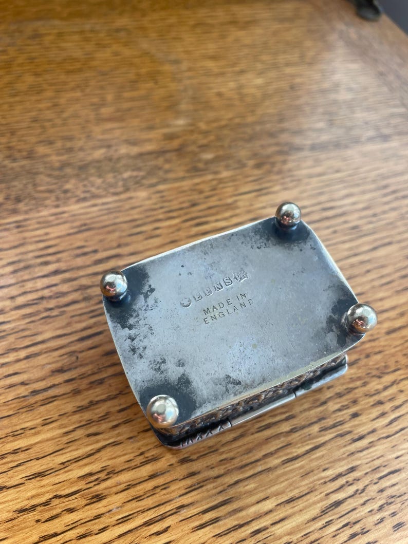 Op de afbeelding: Een kleine, rechthoekige zilveren doos met afgeronde voetjes. De doos heeft een scharnierend deksel en is gemarkeerd met "LENS" en "MADE IN ENGLAND". De doos staat op een houten oppervlak.