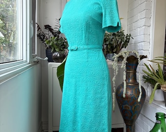 May include: A turquoise vintage dress with a textured floral pattern. The dress features short sleeves, a fitted bodice, and a matching belt. The dress is displayed on a mannequin.