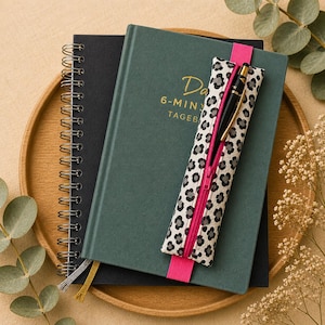 Könnte beinhalten: Ein schwarzes Spiralnotizbuch und ein dunkelgrünes Hardcover-Journal mit goldenen Buchstaben werden auf einem Holztablett präsentiert. Ein Stift befindet sich in einem Stifthalter mit Leopardenmuster und einem rosa Reißverschluss. Ein rosa Band dient als Lesezeichen.