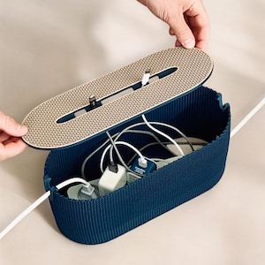 May include: A navy blue cable tidy with a textured lid and a beige patterned surface. The open container reveals multiple white cables, plugs, and chargers. The lid is being lifted to show the contents.