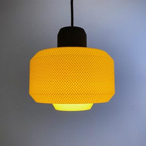 Puede incluir: Lámpara colgante amarilla moderna con una pantalla texturizada en forma de panal. La lámpara tiene una parte superior negra y un cable negro. La pantalla es cilíndrica con una base ligeramente ensanchada, emitiendo un brillo cálido.