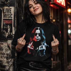 May include: A black short-sleeved t-shirt featuring a graphic print of a person with long hair and round sunglasses, wearing multiple rings and making a middle finger gesture with both hands, rendered in contrasting red and blue color tones.