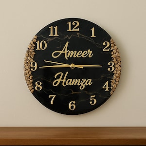 May include: Black and gold clock with a marble design. The clock face has gold numbers and hands, and the words "Ameer Hamza" are written in gold script.