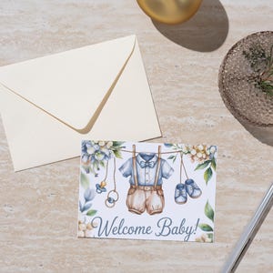 Può includere: Biglietto di auguri per baby shower con un'illustrazione ad acquerello di vestiti per bambini appesi a un filo. Il biglietto dice "Welcome Baby!" ed è accompagnato da una busta color crema e una penna d'argento.