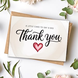 Puede incluir: Una tarjeta de agradecimiento con un fondo de acuarela y un sobre marrón. La tarjeta presenta las palabras "Thank you" en una elegante escritura negra, con un corazón rojo debajo. El texto "A little card to say a big" está encima del texto principal.