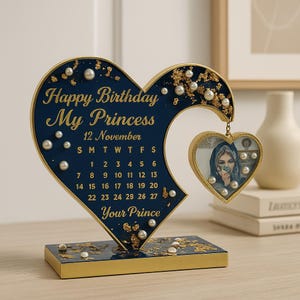 May include: A blue and gold heart-shaped desktop calendar with a smaller heart-shaped photo frame hanging from the top. The calendar features the text "Happy Birthday My Princess" and a calendar for November. The photo frame has a picture of a person wearing a headscarf. The base of the calendar is gold.