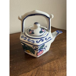 Puede incluir: Una tetera de cerámica blanca con cuerpo hexagonal y asa alta arqueada. La tetera presenta bordes con volutas azules pintadas a mano y motivos frutales coloridos que incluyen detalles en rosa y verde tanto en la tapa como en el cuerpo.