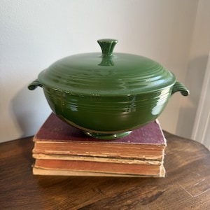 May include: A forest green ceramic covered casserole dish with a glossy finish, featuring concentric ring patterns on the lid and body, a central knob handle on the lid, and two small, curved side handles extending from the bowl's rim.