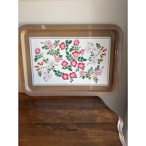 Puede incluir: Una bandeja rectangular con un borde de color beige sólido y un panel central blanco. El centro presenta una ilustración botánica de rosas silvestres rosas y blancas con hojas y tallos verdes dispuestos sobre la superficie.
