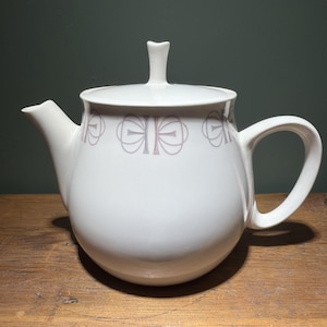 May include: A white ceramic teapot featuring a rounded body, a curved handle, and a spout. The upper portion of the teapot is decorated with a repeating pattern of thin, light gray and muted pink geometric bow-like shapes encircling the vessel.
