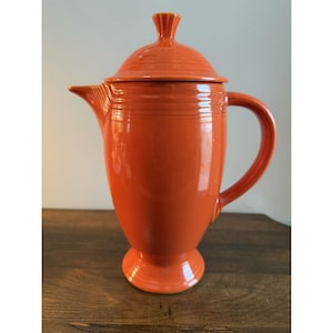 May include: A bright orange ceramic coffee pot with a matching lid, featuring a tall, tapered body, a pedestal base, a curved handle, and a pointed spout. The exterior surface is decorated with horizontal ringed ridges near the top rim.