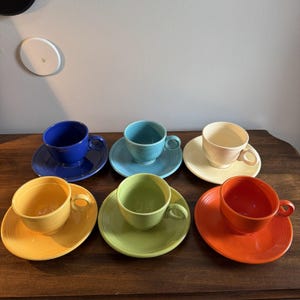 Puede incluir: Un juego de seis tazas de té de cerámica con sus platos a juego en colores vibrantes que incluyen azul cobalto, turquesa, crema, amarillo, verde claro y naranja. Cada taza tiene un asa redondeada y detalles de anillos concéntricos.
