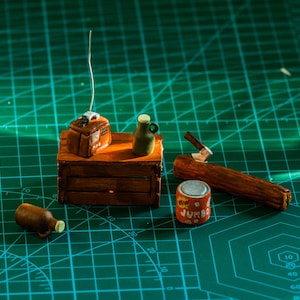 Pode incluir: Uma cena em miniatura com uma caixa de madeira, um rádio, uma garrafa, uma lata e um tronco de madeira. A cena está situada em um tapete de corte verde.