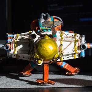 May include: A model of a spaceship with a gold dome, white and brown body, and blue and orange accents. The spaceship has two cannons on the sides and is standing on a black surface.