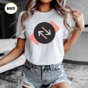 Könnte beinhalten: Weißes T-Shirt mit einem schwarzen Kreis, der zwei weiße Pfeile enthält, die in entgegengesetzte Richtungen zeigen. Der Kreis ist von einer rosa Form umgeben.