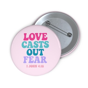 Könnte beinhalten: Zwei runde, hellrosa Knöpfe. Ein Knopf zeigt den Text "LOVE CASTS OUT FEAR" in Pink, Türkis, Blau und Lila. Der Text "1 JOHN 4:18" steht darunter. Der andere Knopf ist silberfarben.