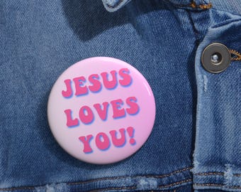 May include: A round, pink button with the text "JESUS LOVES YOU!" in blue and pink lettering. The button is pinned to a blue denim jacket.