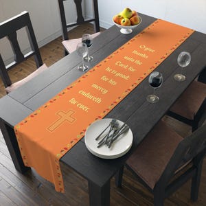 Op de afbeelding: Oranje tafelloper met een kruisontwerp en de tekst "O give thanks unto the Lord: for he is good: for his mercy endureth for ever." De loper ligt op een donkere houten tafel, gedekt met borden, bestek en wijnglazen.