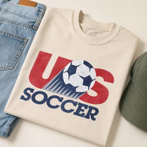 May include: A cream-colored crew neck t-shirt featuring a distressed graphic with the red letters U and S, a blue and white soccer ball, and the blue text SOCCER below. The design includes a motion trail effect behind the soccer ball.
