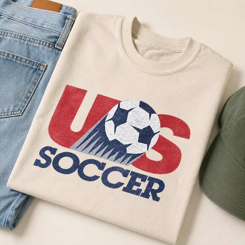May include: A cream-colored crew neck t-shirt featuring a distressed graphic with the red letters U and S, a blue and white soccer ball, and the blue text SOCCER below. The design includes a motion trail effect behind the soccer ball.