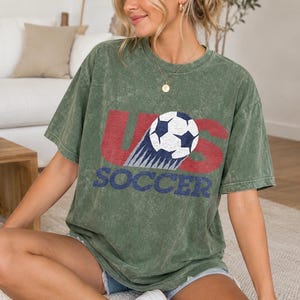 May include: An olive green mineral-washed short-sleeve t-shirt featuring a graphic print with the red letters US and the blue word SOCCER. A white and black soccer ball graphic is positioned over the letter S, showing a motion trail effect.