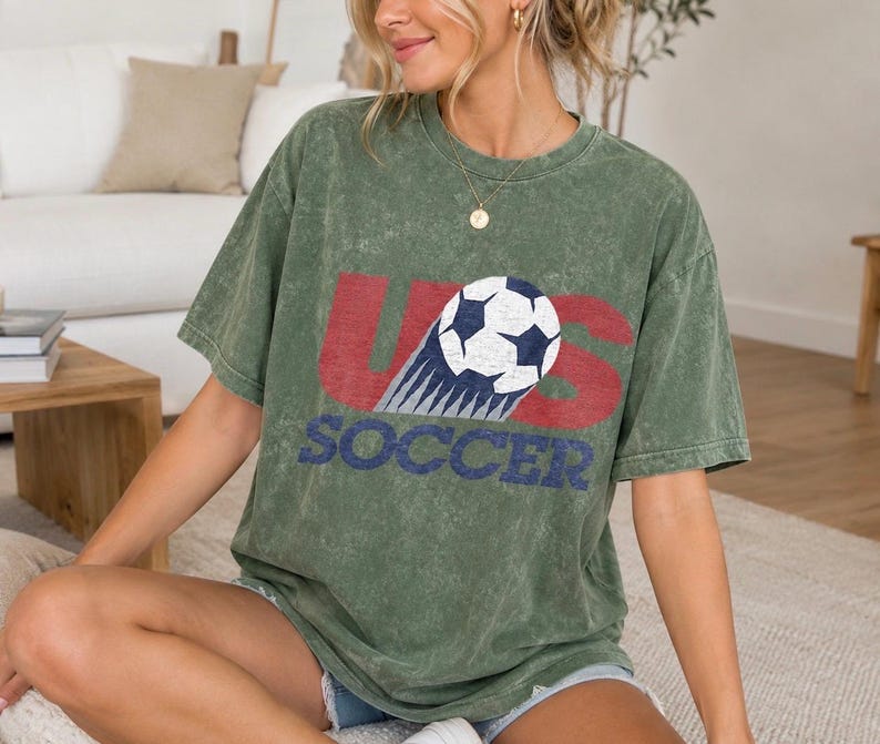 May include: An olive green mineral-washed short-sleeve t-shirt featuring a graphic print with the red letters US and the blue word SOCCER. A white and black soccer ball graphic is positioned over the letter S, showing a motion trail effect.
