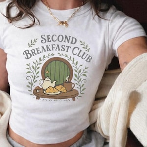 Puede incluir: Camiseta blanca con un diseño gráfico que dice "SECOND BREAKFAST CLUB". El diseño incluye una imagen de dibujos animados de una mesa con una tetera, cruasanes y una taza de café frente a una puerta verde, rodeada de hojas verdes.