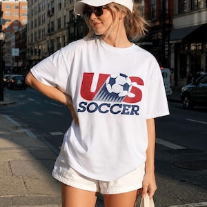 May include: A white short-sleeve t-shirt featuring a graphic print with the red letters US, a black and white soccer ball, and the blue text SOCCER below. The shirt is paired with a white baseball cap and white shorts.