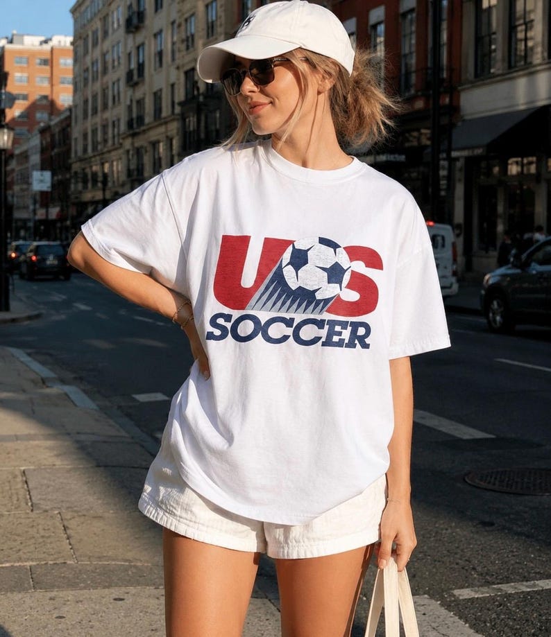 May include: A white short-sleeve t-shirt featuring a graphic print with the red letters US, a black and white soccer ball, and the blue text SOCCER below. The shirt is paired with a white baseball cap and white shorts.