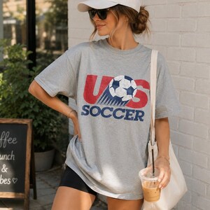 May include: A heather gray short-sleeve t-shirt featuring a distressed graphic print that reads US SOCCER in red and blue lettering, with a soccer ball replacing the letter O. The shirt has a crew neckline and a loose, relaxed fit design.