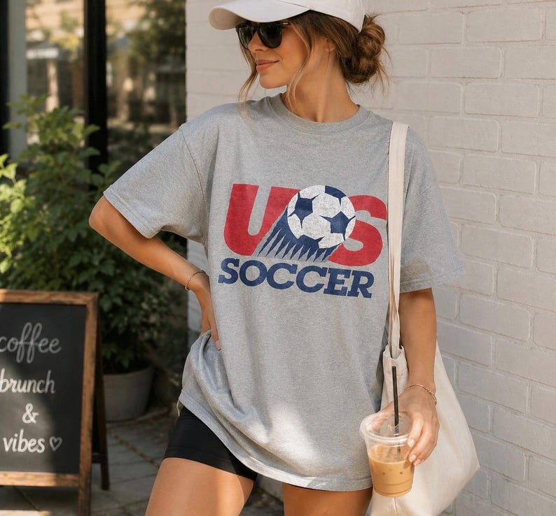 May include: A heather gray short-sleeve t-shirt featuring a distressed graphic print that reads US SOCCER in red and blue lettering, with a soccer ball replacing the letter O. The shirt has a crew neckline and a loose, relaxed fit design.