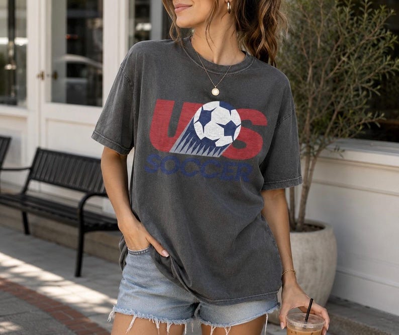 May include: A charcoal grey short-sleeve t-shirt featuring a graphic print with the red letters US and the blue word SOCCER. A black and white soccer ball graphic is positioned between the letters, showing a motion trail effect on the shirt.