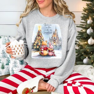 A woman wearing a gray sweatshirt with a Christmas-themed graphic and holding a mug of hot chocolate is sitting in front of a Christmas tree.
